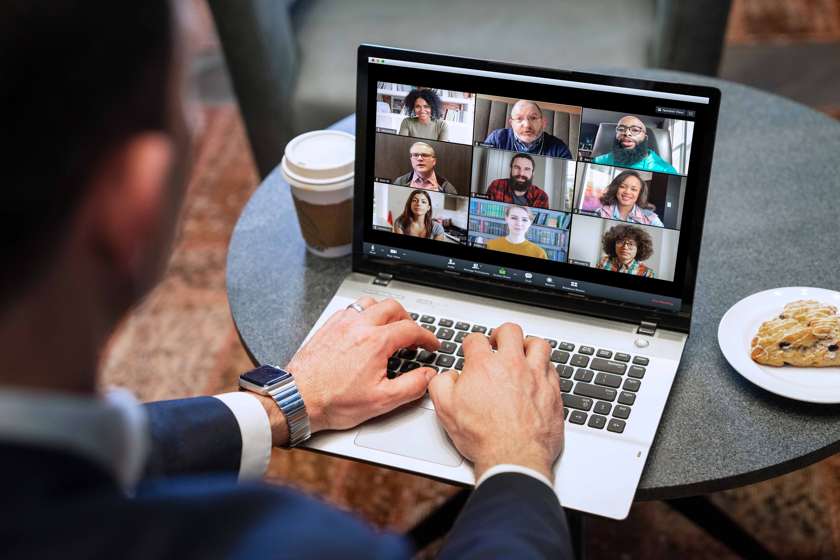 A man dials into a video chat with colleagues on his laptop while having coffee and a scone