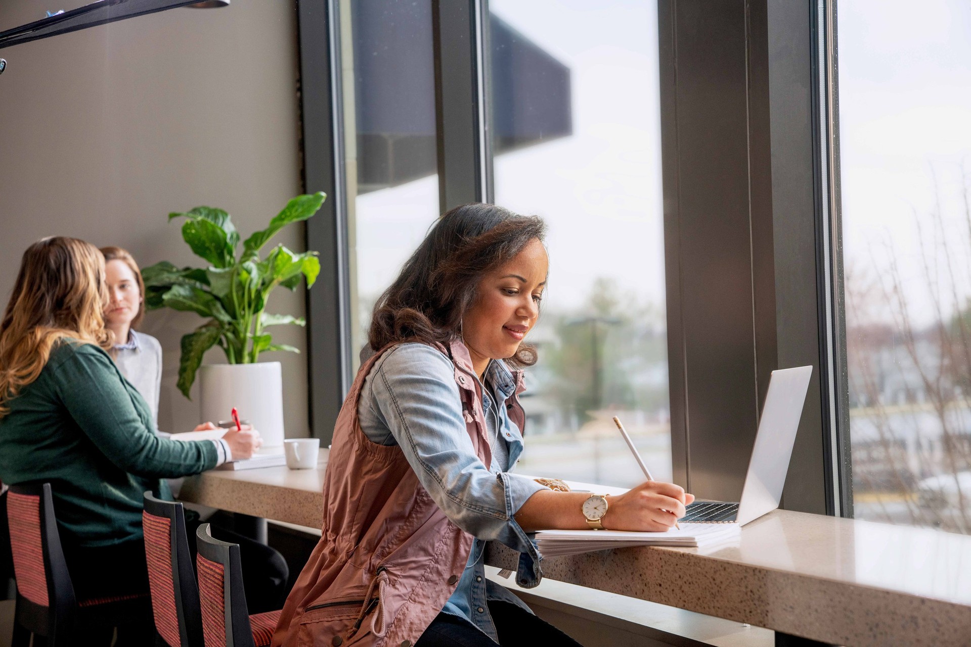 A woman writes on a notepad while sitting in a cafe lounge and working on her laptop