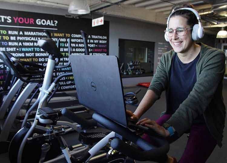 A woman on an exercise bike working on her laptop and listing to her headphones